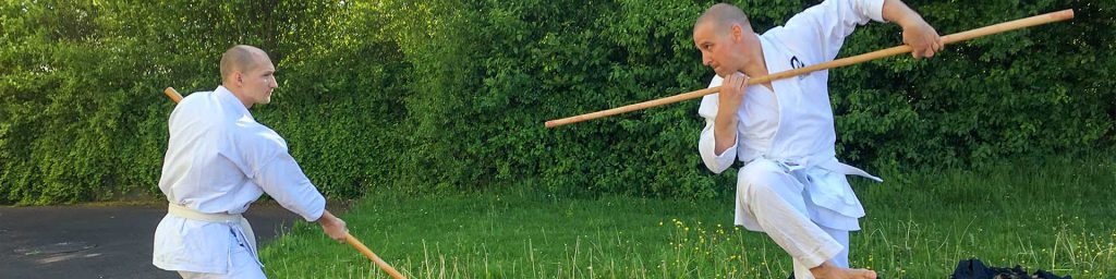 Zwei Männer üben Kobudo Techniken mit dem Bo auf einer Wiese.